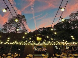 An outdoor seating area with string lights and a neon sign for 'Tad's Bar & Grill.' - Cabin on The Hill at 4 Guys RV Park Campground (Stanton)