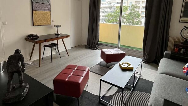 Un salon moderne comprenant un coin repas, un canapé, un pouf rouge, une petite table avec un saladier et une statue.