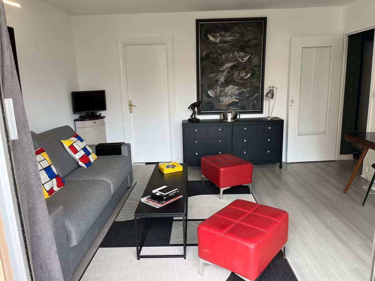 Una sala de estar con un sofá gris, un otoman rojo, una mesa de centro negra y un televisor negro sobre un soporte.