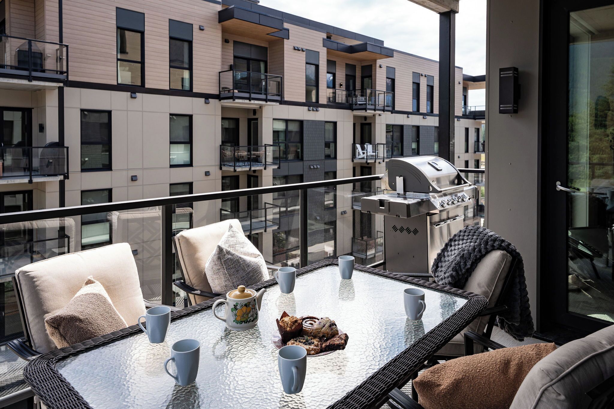A balcony with a glass table, chairs, a grill, and a view of a building with multiple balconies.