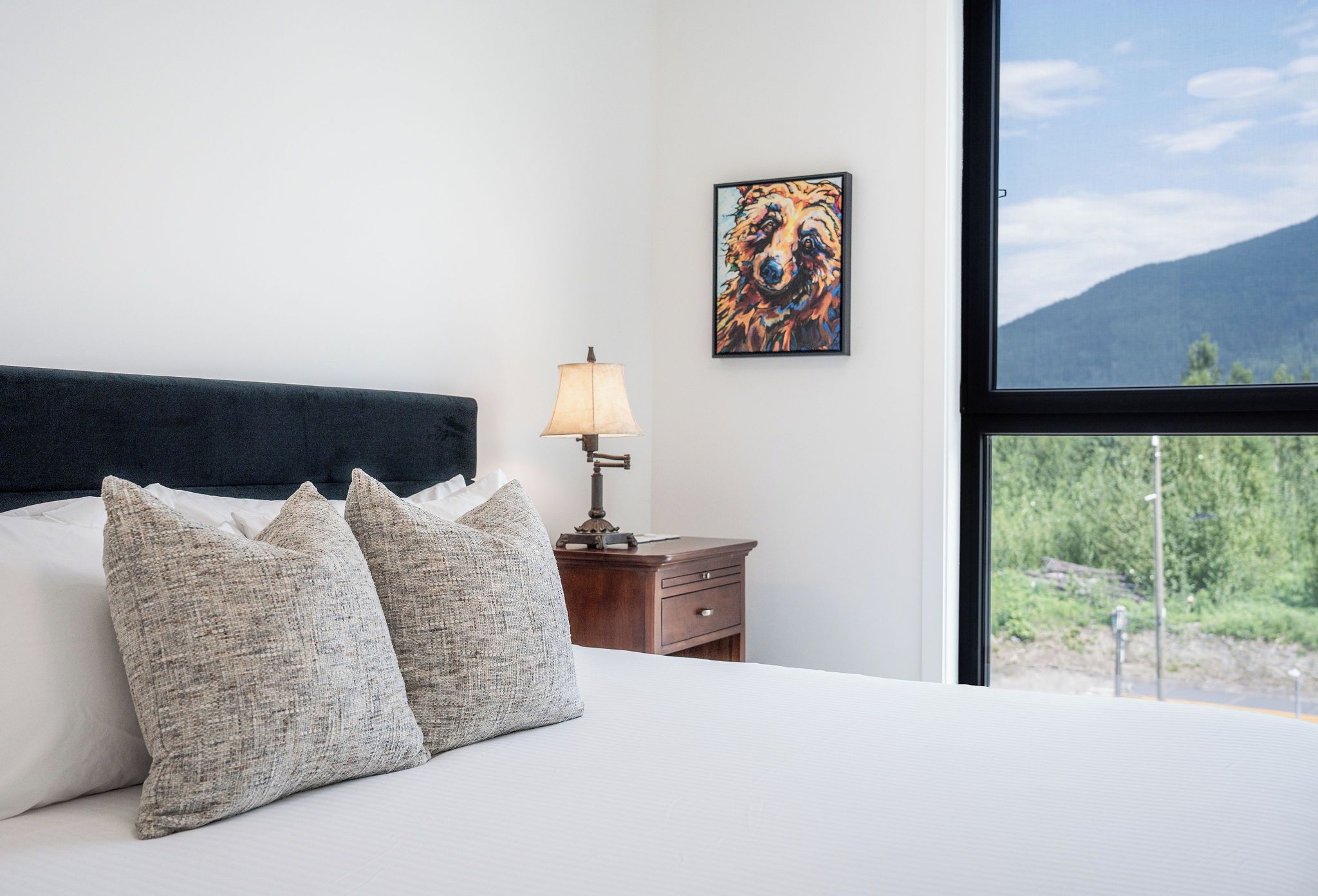 A bedroom with a bed, pillows, a nightstand with a lamp, a painting, and a window with a mountain view.