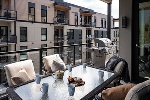 A balcony with a glass table, chairs, a grill, and a view of a building with multiple balconies. - Hot Sauce Hideaway -Revelstoke Vacations (Revelstoke)