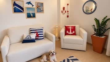 Sala de estar com sofá, poltrona e um cão pequeno. Decorações nas paredes incluem quadros e um espelho.