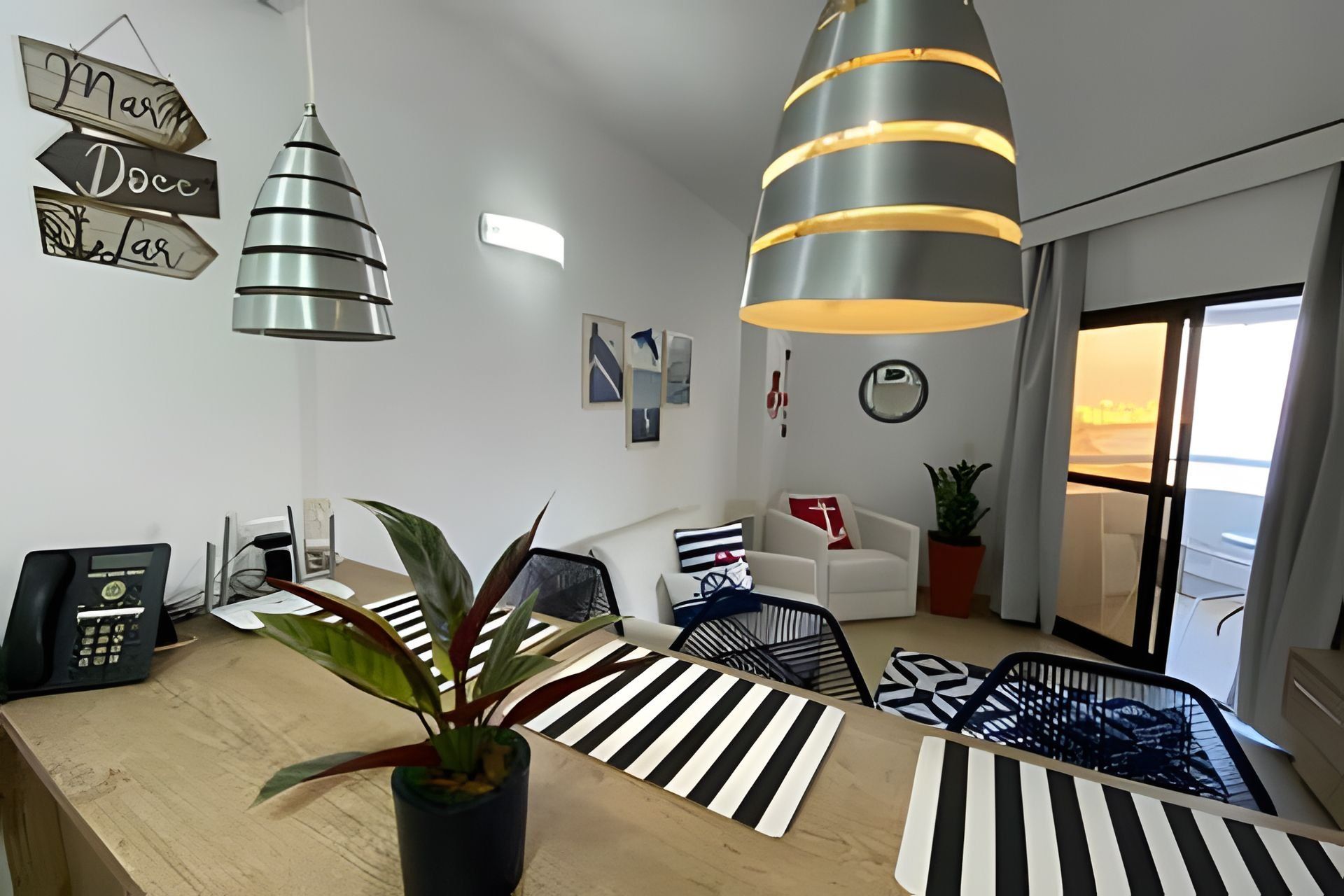 A modern living room with a wooden dining table, striped rugs, and a potted plant. There are two hanging pendant lights and a large window with a view.