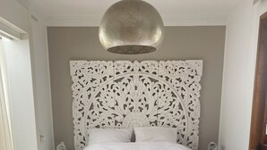 A bedroom with a large bed, a decorative headboard, a hanging light fixture, and a wooden bench. - Dune Villa with a View | Greenhouse, Piano, and Beach Nearby (Ouddorp)