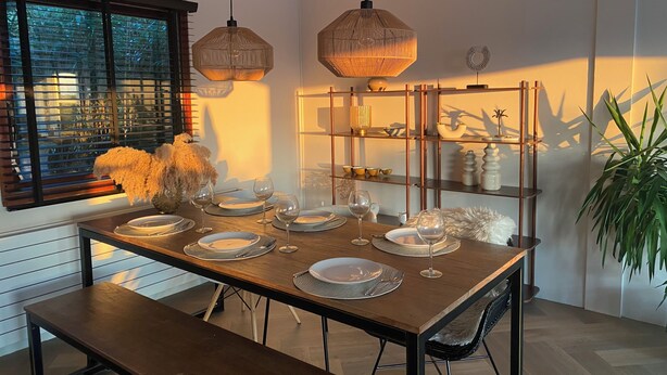 A dining area with a wooden table set for four, two hanging pendant lights, a glass shelf with decorative items, and a large window with blinds. - Dune Villa with a View | Greenhouse, Piano, and Beach Nearby (Ouddorp)