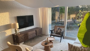 A modern living room with a flat-screen TV, a wooden cabinet, two beige armchairs, a round coffee table, and a sofa. - Dune Villa with a View | Greenhouse, Piano, and Beach Nearby (Ouddorp)