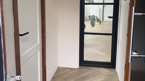 A hallway with a wooden floor, a door with a black handle, a ceiling light fixture, and a view through a glass door. - Dune Villa with a View | Greenhouse, Piano, and Beach Nearby (Ouddorp)