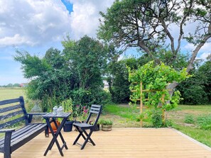 Eine Holzterrasse mit Bank und Tisch, umgeben von Grünpflanzen und einem mit Weinreben bewachsenen Spalier.