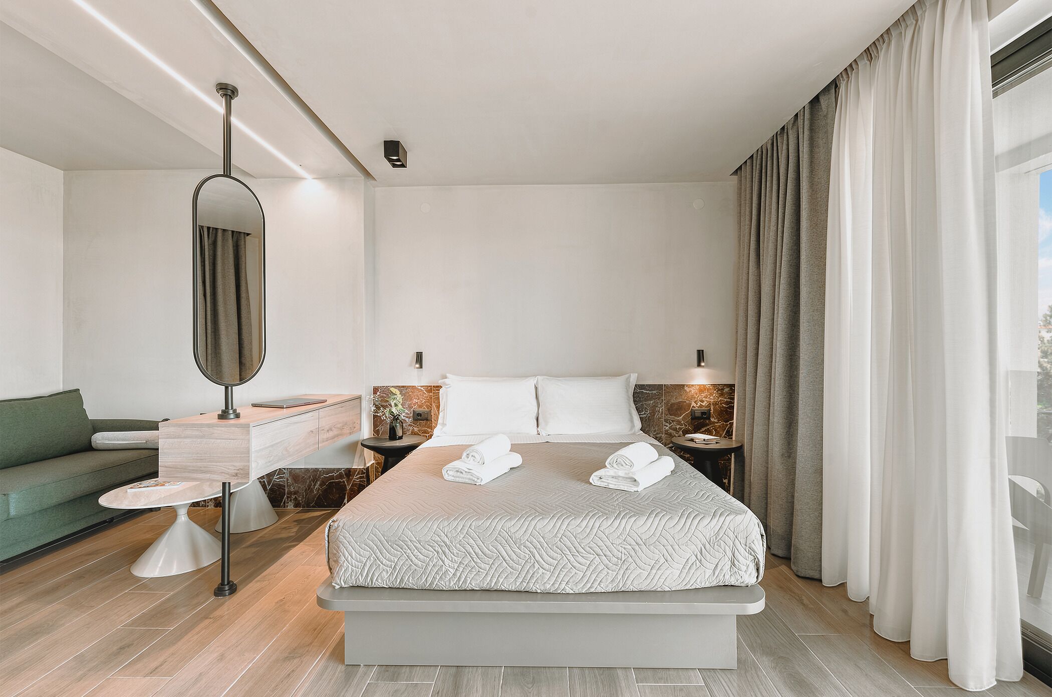 Ein modernes Hotelzimmer mit einem großen Bett, einem Nachttisch, einem Spiegel, einem grünen Sofa und Blick ins Freie.