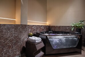 A modern bathroom with a marble wall, a heated floor, a black bathtub, and a potted plant.