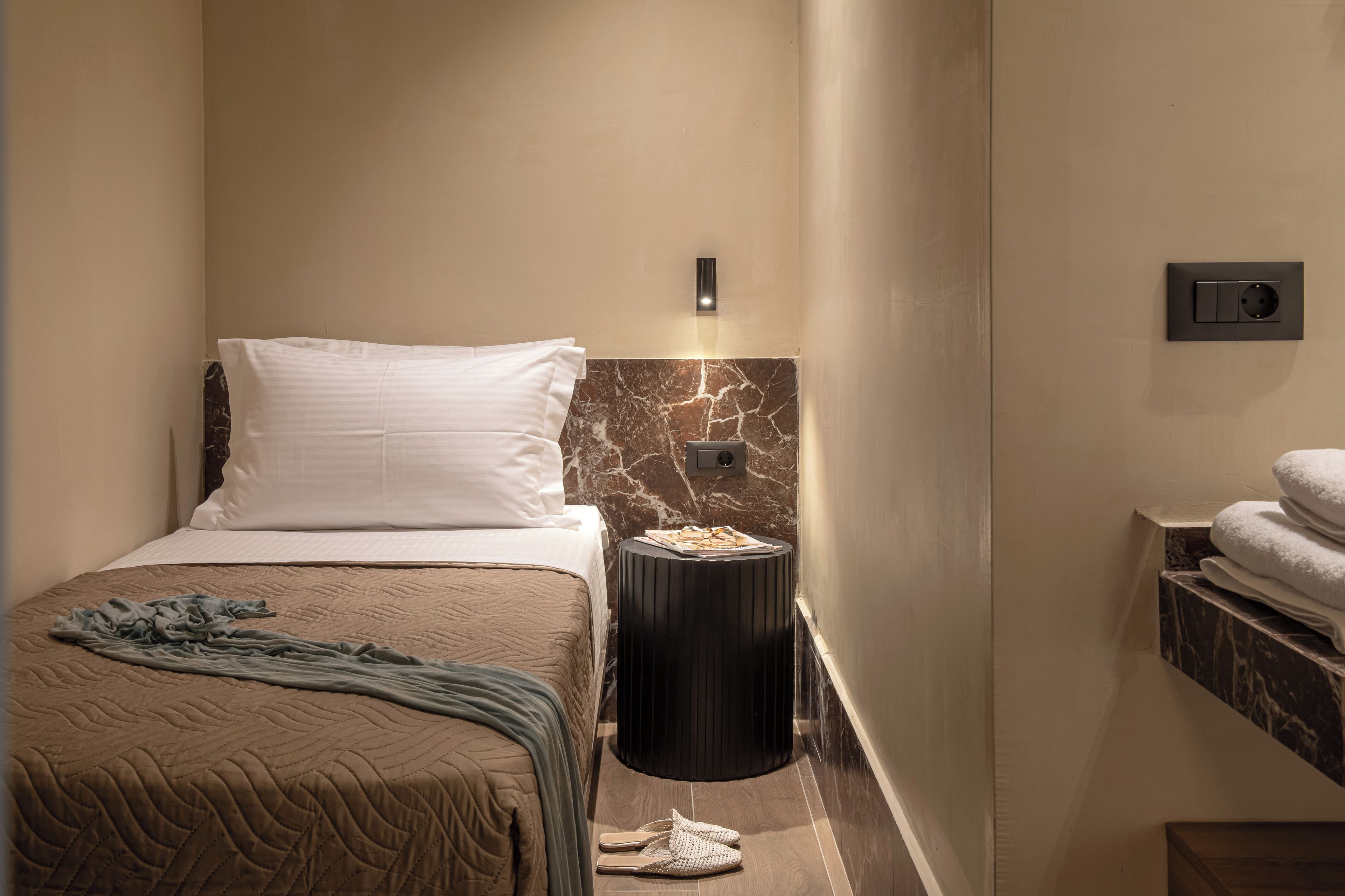 A hotel room with a bed, a nightstand, a marble wall, a towel rack, and a power outlet.