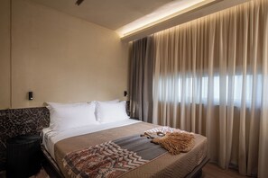 A hotel room with a bed, a nightstand, a wall-mounted lamp, and a window with curtains.
