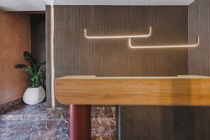 A reception desk with a wooden finish, a potted plant, and a wall with vertical lines and a neon light. - MAHA Her City Living by Semavi (Heraklion)