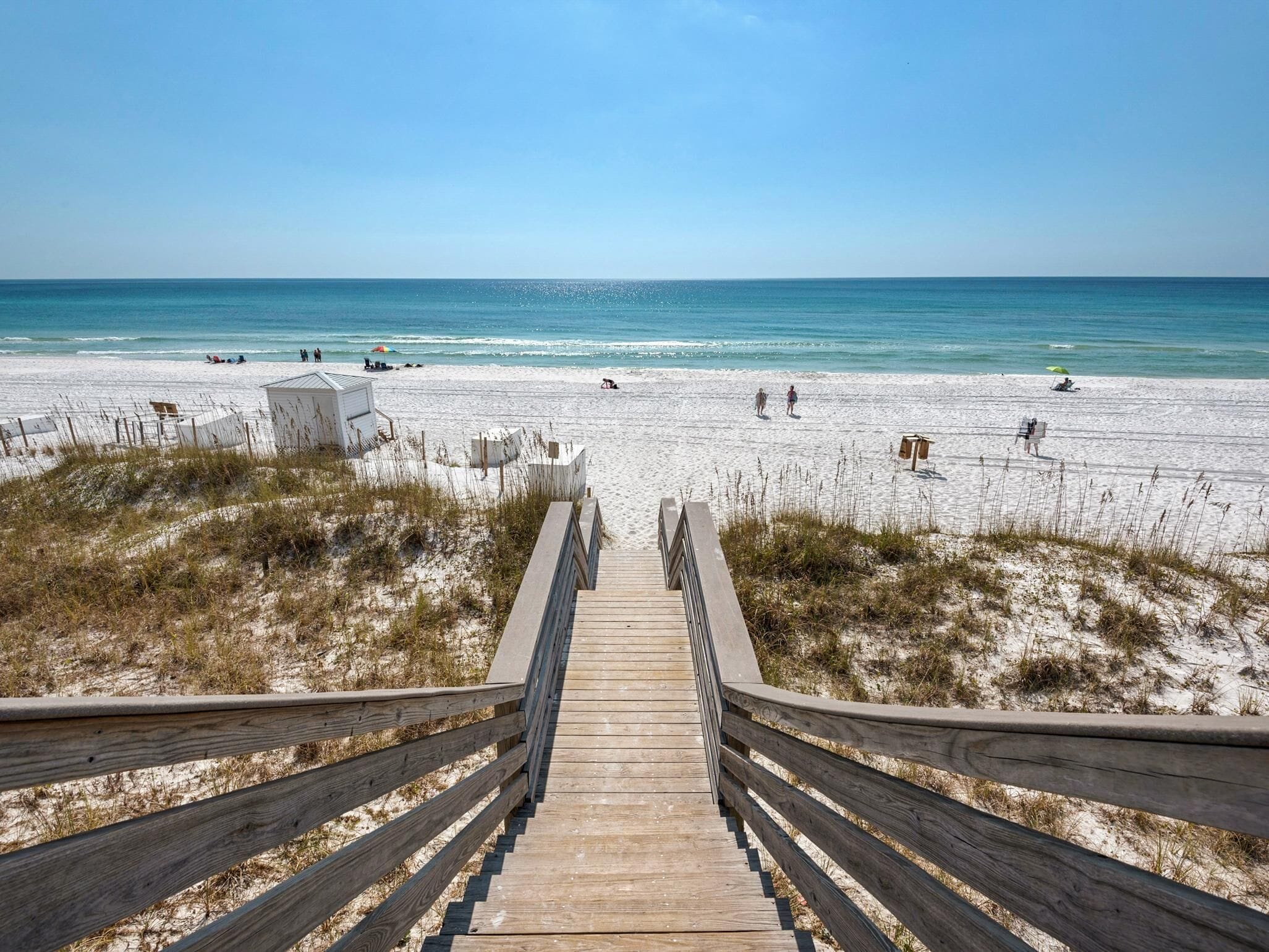 Un escalier en bois mène à une plage de sable blanc, sous un ciel bleu azur.