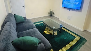 A living room with a sofa, a glass coffee table, a green patterned rug, a television on the wall, and a ceiling light.