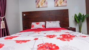 Quarto com cabeceira de madeira, colcha vermelha e branca com motivos florais, duas almofadas, mesa de cabeceira, controle remoto e um vaso com planta.