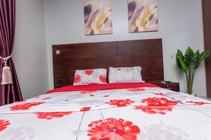 Ein Schlafzimmer mit einem hölzernen Kopfteil, einer rot-weißen blumigen Tagesdecke, zwei Kissen, einem Nachttisch, einer Fernbedienung und einem Blumentopf.