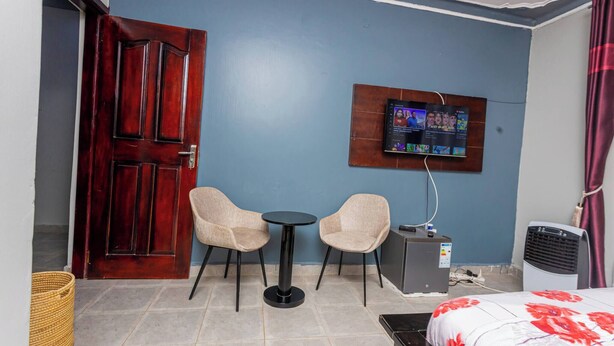 Quarto de hotel com porta de madeira, duas cadeiras bege, uma mesinha redonda, televisão na parede, aquecedor e uma cama com colcha florida.