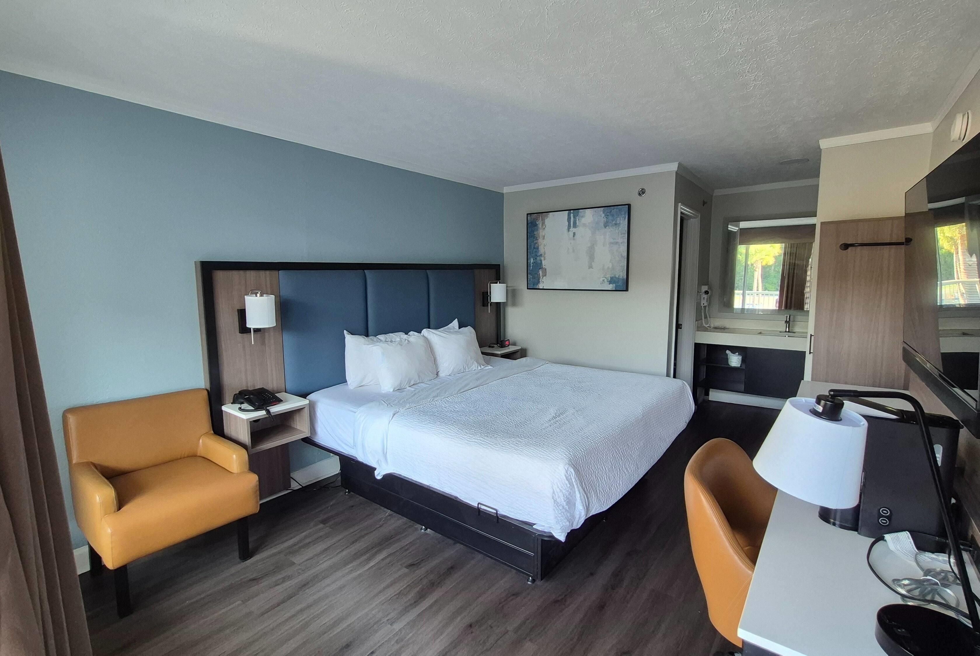Quarto de hotel com cama, escrivaninha, cadeira, mesa de cabeceira e um quadro na parede.
