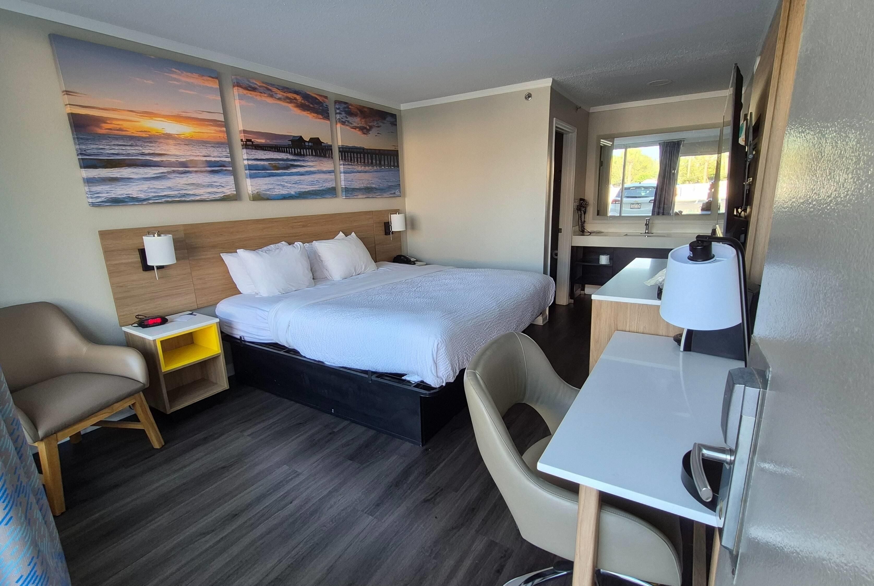 A hotel room with a bed, a desk, a chair, a nightstand, and a painting of a sunset over the ocean.