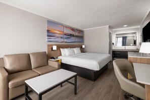 A hotel room with a bed, a sofa, a desk, and a chair. There are two wall-mounted lamps, a telephone, and a painting of a sunset. - Days Inn By Wyndham Pawleys Island (Pawleys Island)