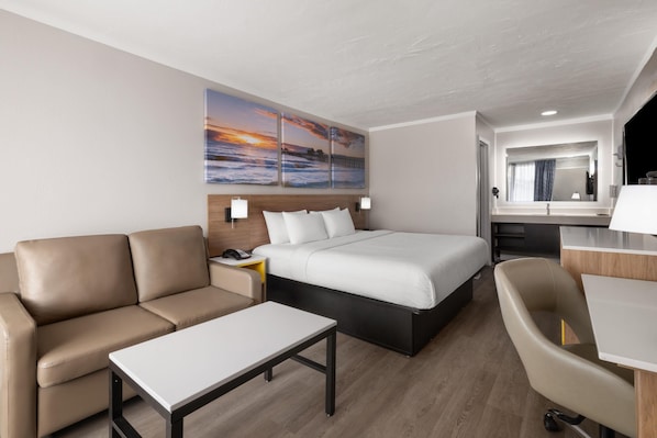 A hotel room with a bed, a sofa, a desk, and a chair. There are two wall-mounted lamps, a telephone, and a painting of a sunset.