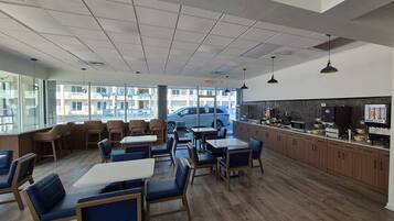 A restaurant with a modern interior, featuring a mix of seating options including tables and booths, a kitchen area with visible appliances, and large windows offering a view of the street.