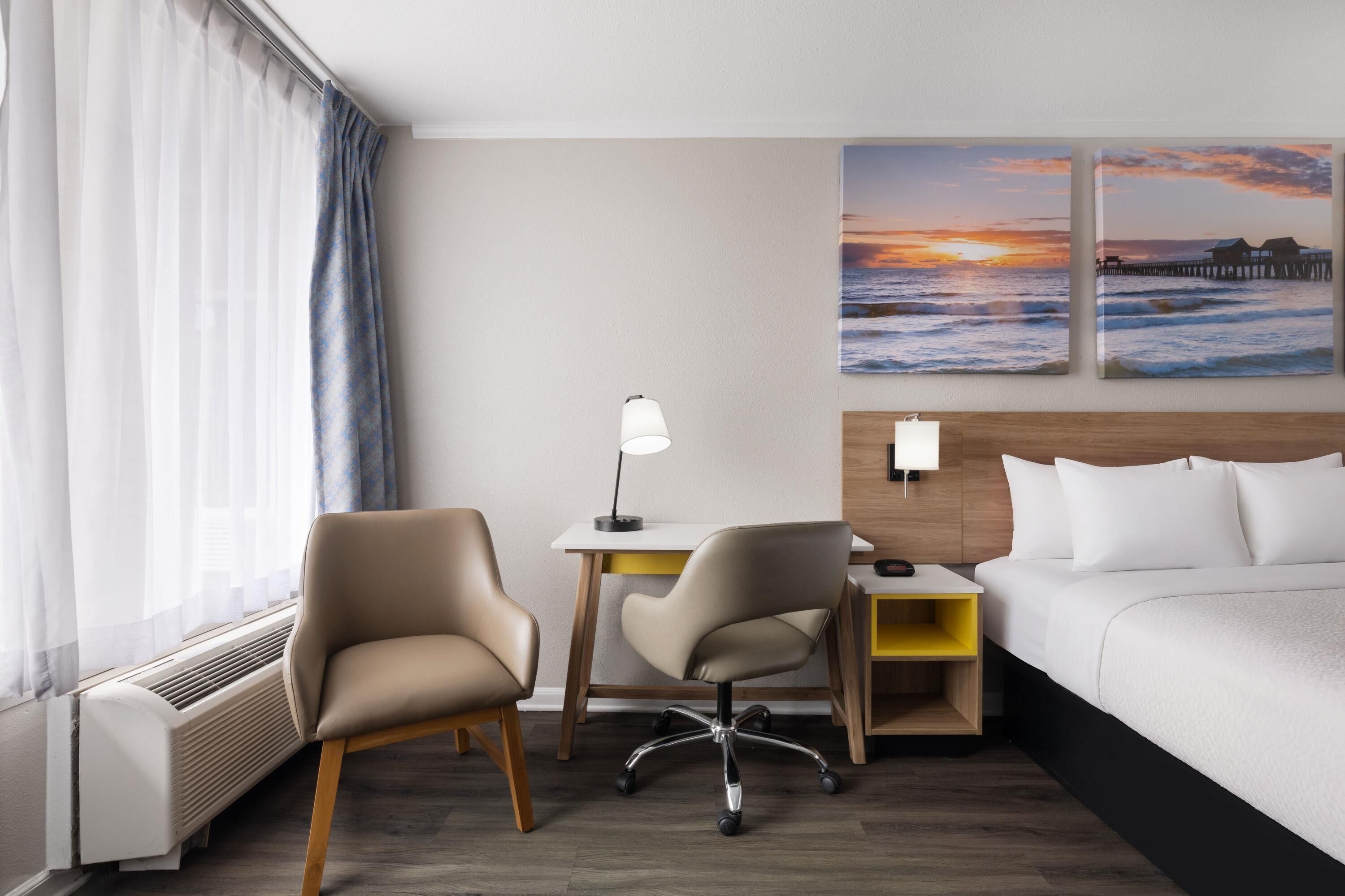 A hotel room with a bed, two chairs, a desk, a lamp, and two wall art pieces depicting a sunset over the ocean.