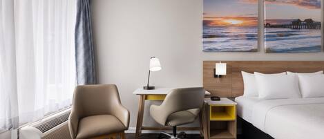 A hotel room with a bed, two chairs, a desk, a lamp, and two wall art pieces depicting a sunset over the ocean.