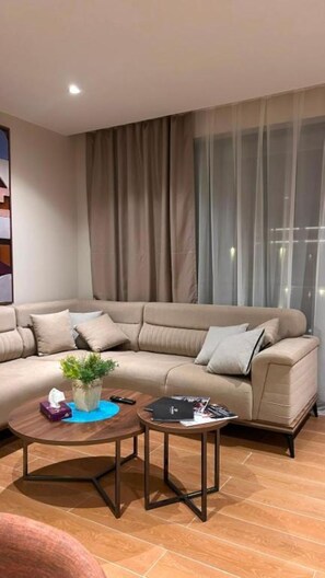 Ein modernes Wohnzimmer mit einem beigefarbenen Ecksofa, zwei Couchtischen und einem großen Fenster mit transparenten Vorhängen.