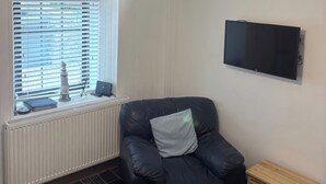 A room with a black leather chair, a small wooden table, a radiator, a window with blinds, and a mounted television. - Viola Cottage (Fraserburgh)