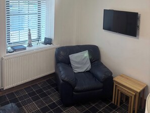 A room with a black leather chair, a small wooden table, a radiator, a window with blinds, and a mounted television. - Viola Cottage (Fraserburgh)