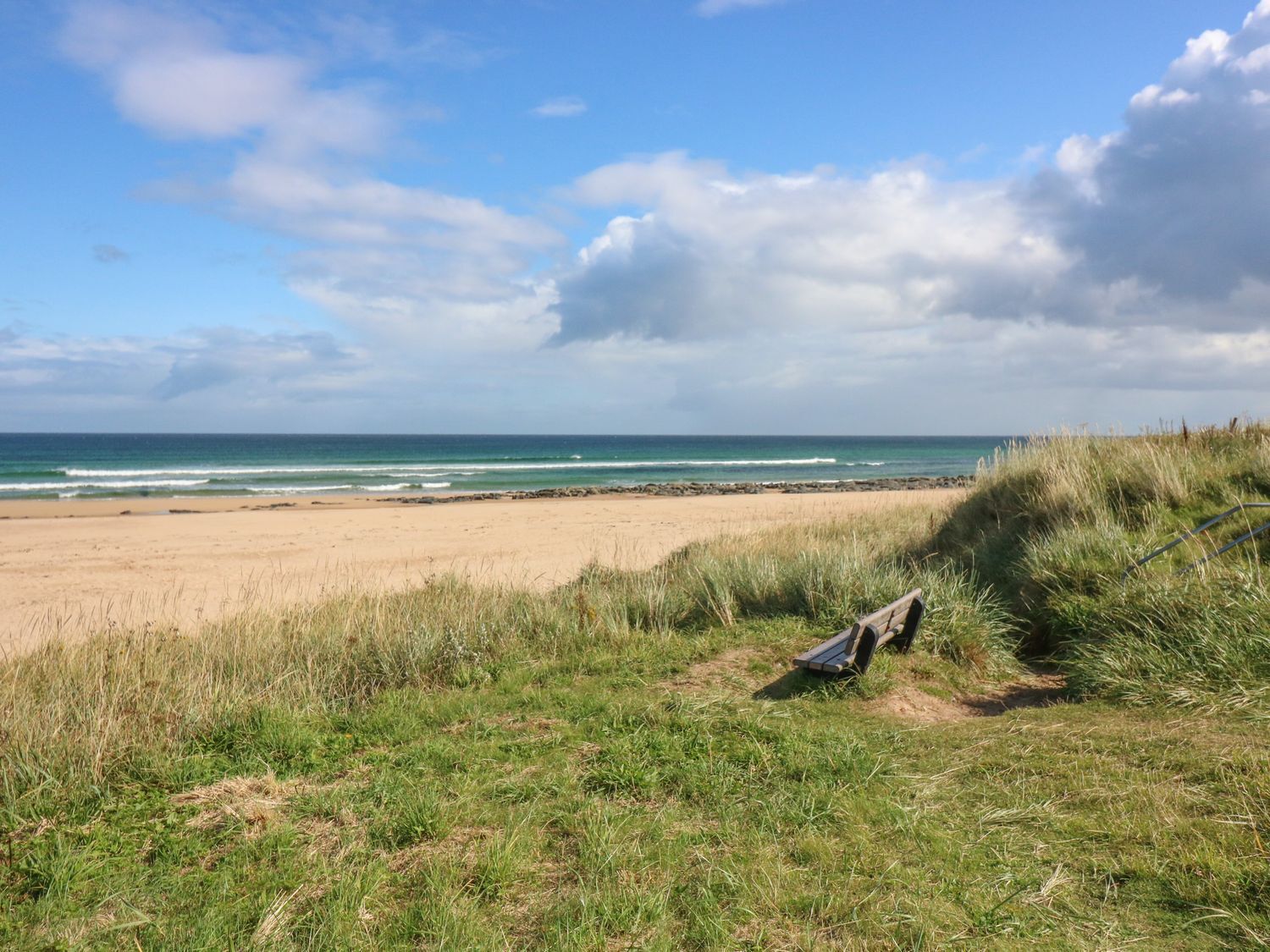 Ein Strand mit sandigem Ufer, grasbewachsenen Dünen und einer Holzbank.