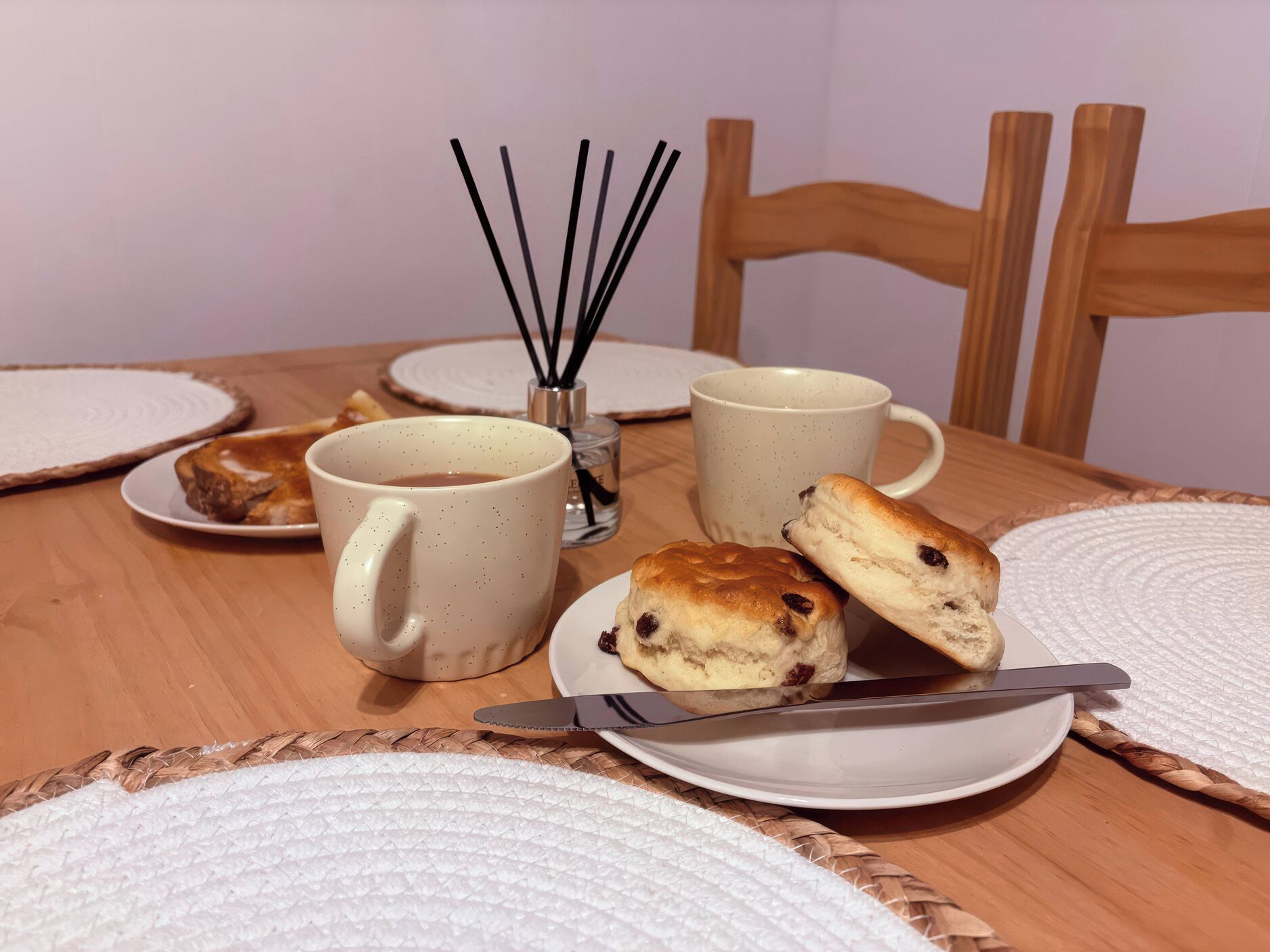 Una mesa con dos tazas, un plato con un pastel, un cuchillo y un difusor con palitos de junco.