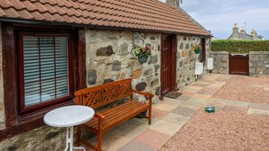 A stone building with a tiled roof, a wooden bench, a small table, and a gate. - Viola Cottage (Fraserburgh)