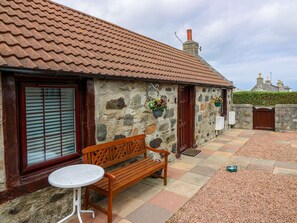 A stone building with a tiled roof, a wooden bench, a small table, and a gate. - Viola Cottage (Fraserburgh)