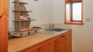 A kitchen with wooden cabinets, a built-in shelf with books, a window, and a sink. - Viola Cottage (Fraserburgh)