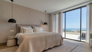 Ein modernes Schlafzimmer mit einem großen Bett, einem Nachttisch, bodentiefen Fenstern mit Blick aufs Meer.