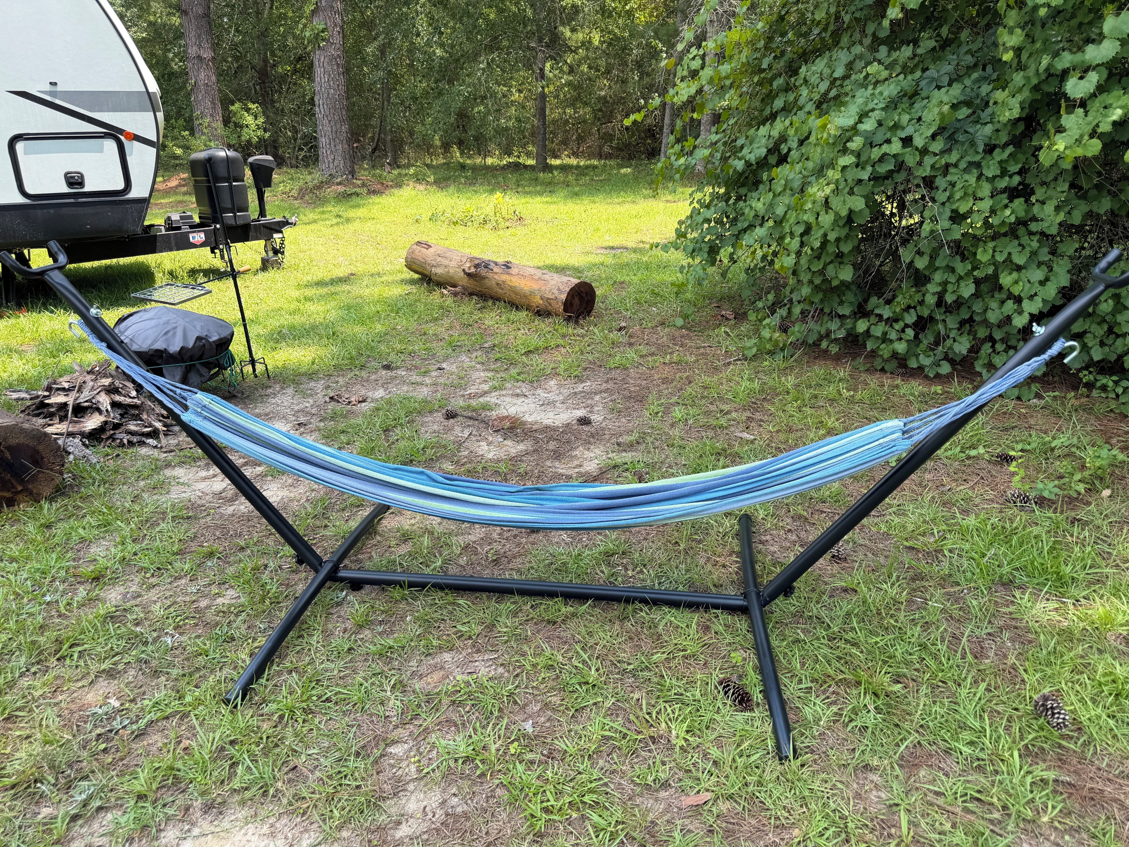 A hammock set up on a stand in a grassy area with a tree and a log nearby.