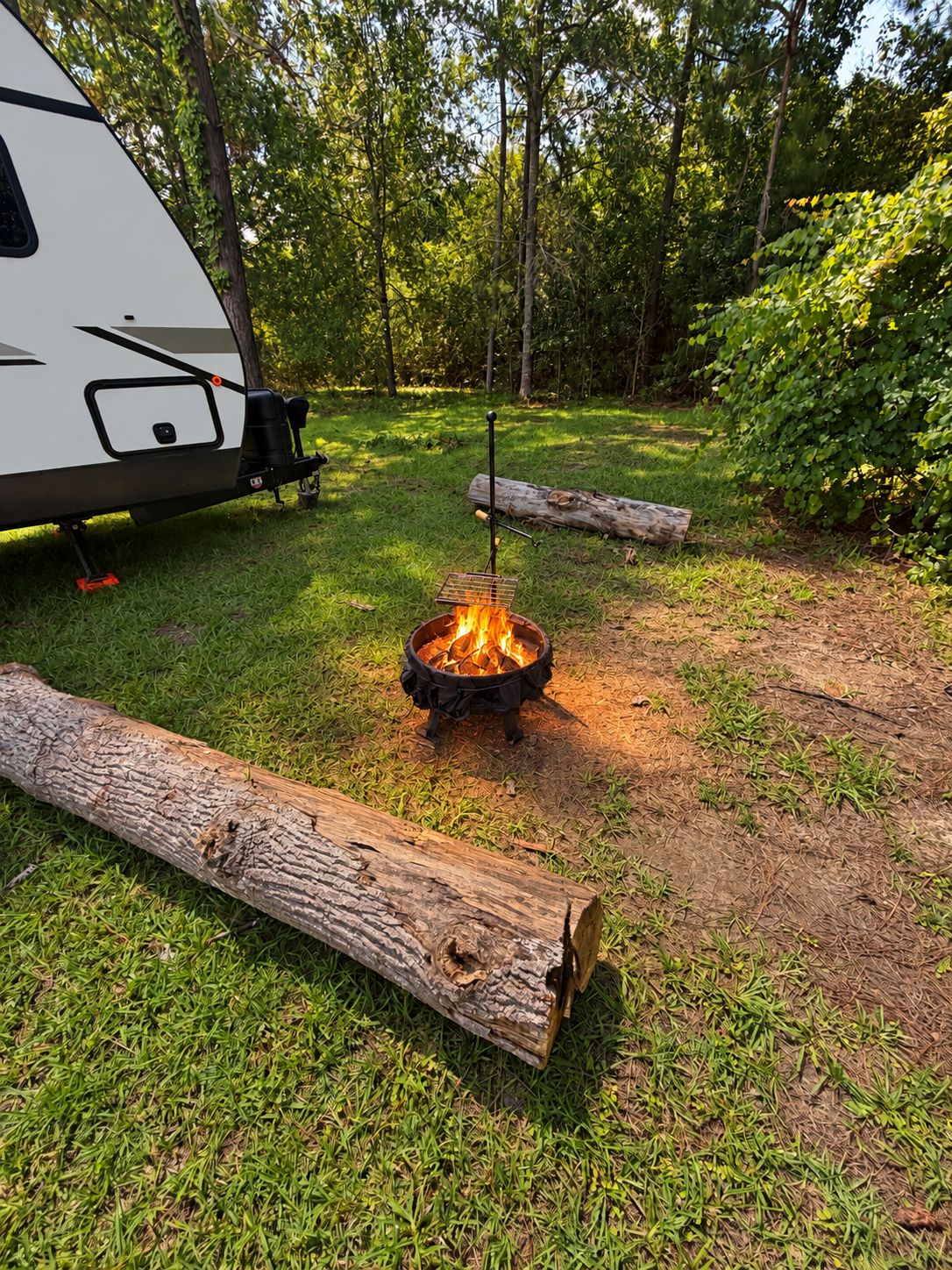 A campsite with a fire pit surrounded by logs, a portable grill, and a white RV.