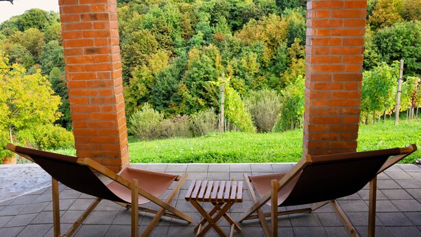 Deux fauteuils de jardin en bois, accompagnés d’une petite table, sont disposés sur une terrasse agrémentée d’un pilier en briques, d’où l’on peut admirer un paysage verdoyant et luxuriant.