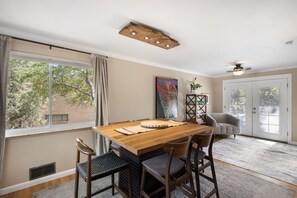 A dining area with a wooden table and chairs, a ceiling light fixture, a painting on the wall, and a view of trees through the window. - THE SUNSET HOUSE 3-Bedroom, 2-Bath House in Central Colorado Springs. (Colorado Springs)