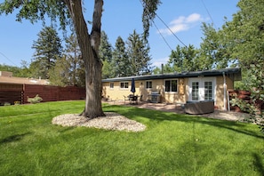 Ein Bungalow mit Terrasse, Grill und einem großen Baum im Garten.
