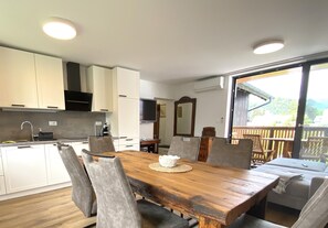 A modern kitchen with white cabinets, a wooden dining table with chairs, and a large window with a view of greenery. - BLED LAKE HOUSE - Apartment Castle (Bled)