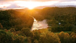 Un río serpentea por un valle boscoso, con un amanecer o atardecer al fondo.