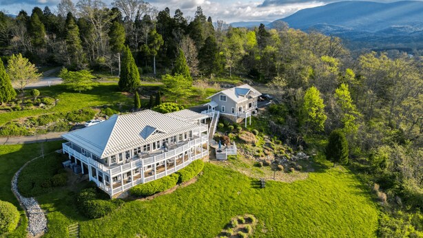 Una casa grande de dos pisos con un porche envolvente y numerosas ventanas, rodeada de abundante vegetación y un paisaje montañoso.