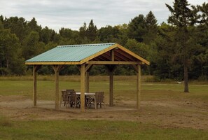 A wooden pavilion with a metal roof, surrounded by chairs and a table, set in a grassy field with trees in the background.