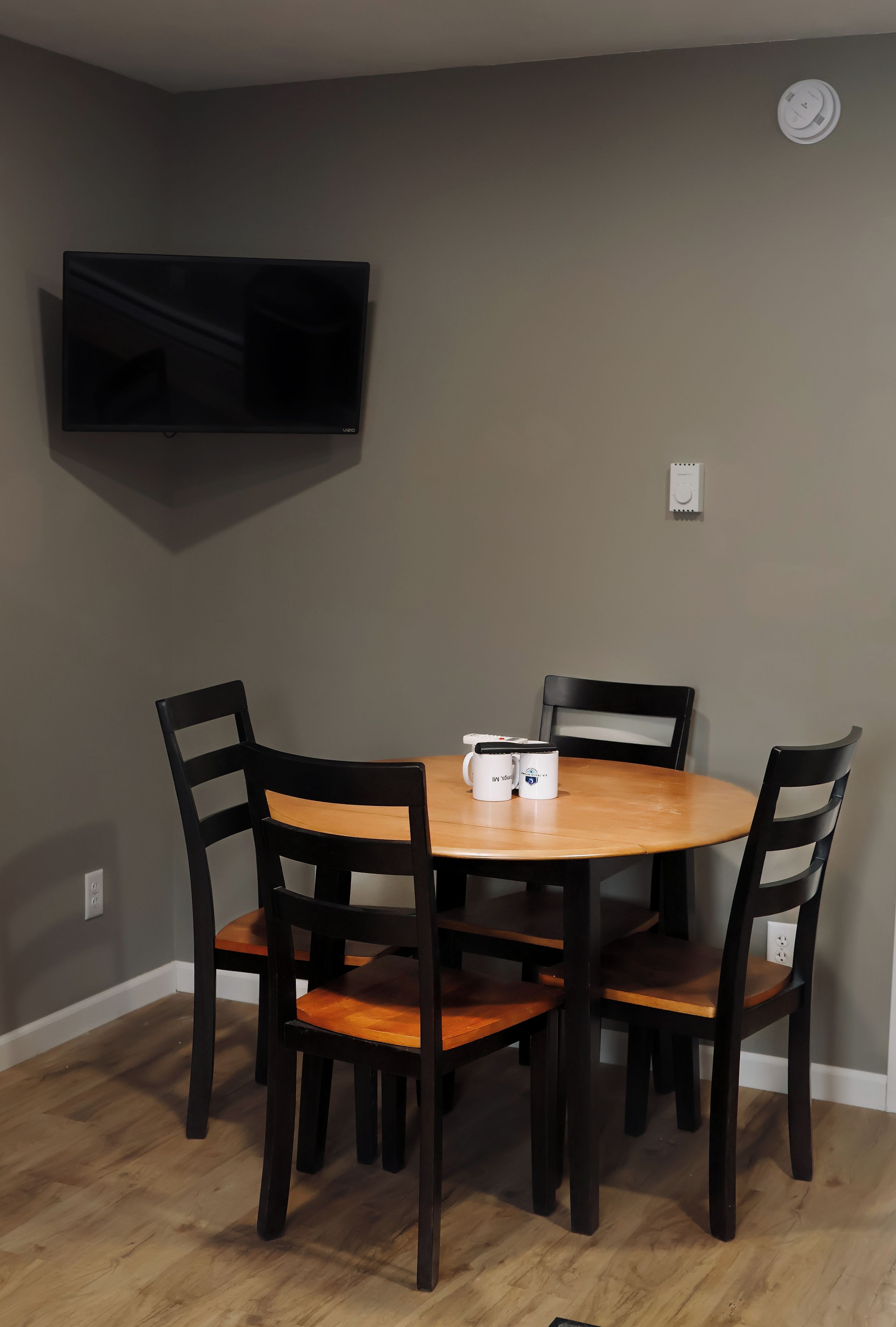 A dining area with a round table, four chairs, a flat-screen TV mounted on the wall, and a power outlet on the wall.