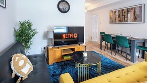 Una sala de estar con un sofá, un sillón amarillo, una mesa de centro, un televisor y un comedor con sillas.